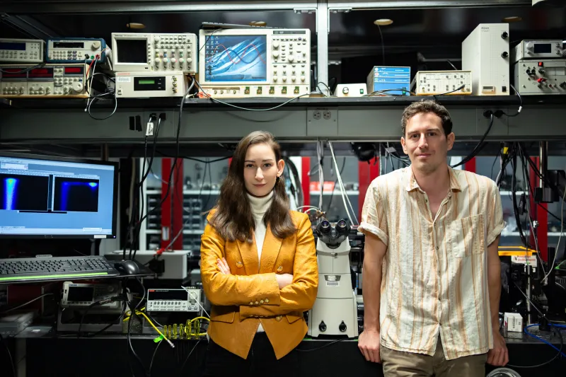 MIT team members Madeleine Laitz (left) and lead author Dane deQuilettes (right) stand in front of multiple computers and machines..