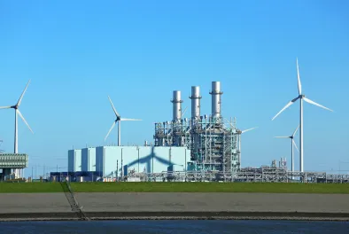 A factory (set against a blue sky) has multiple wind turbines on either side