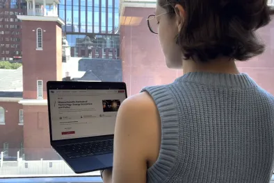 A standing person holds a laptop; the laptop screen features a webpage about MIT and energy policy.